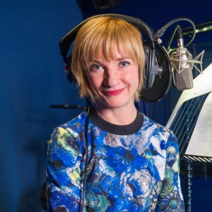 A woman with short blonde hair, wearing a blue shirt, is wearing headphones and sitting close to a microphone in a studio.