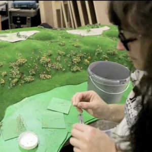 A woman with dark brown hair sitting in front of a green board, making something.